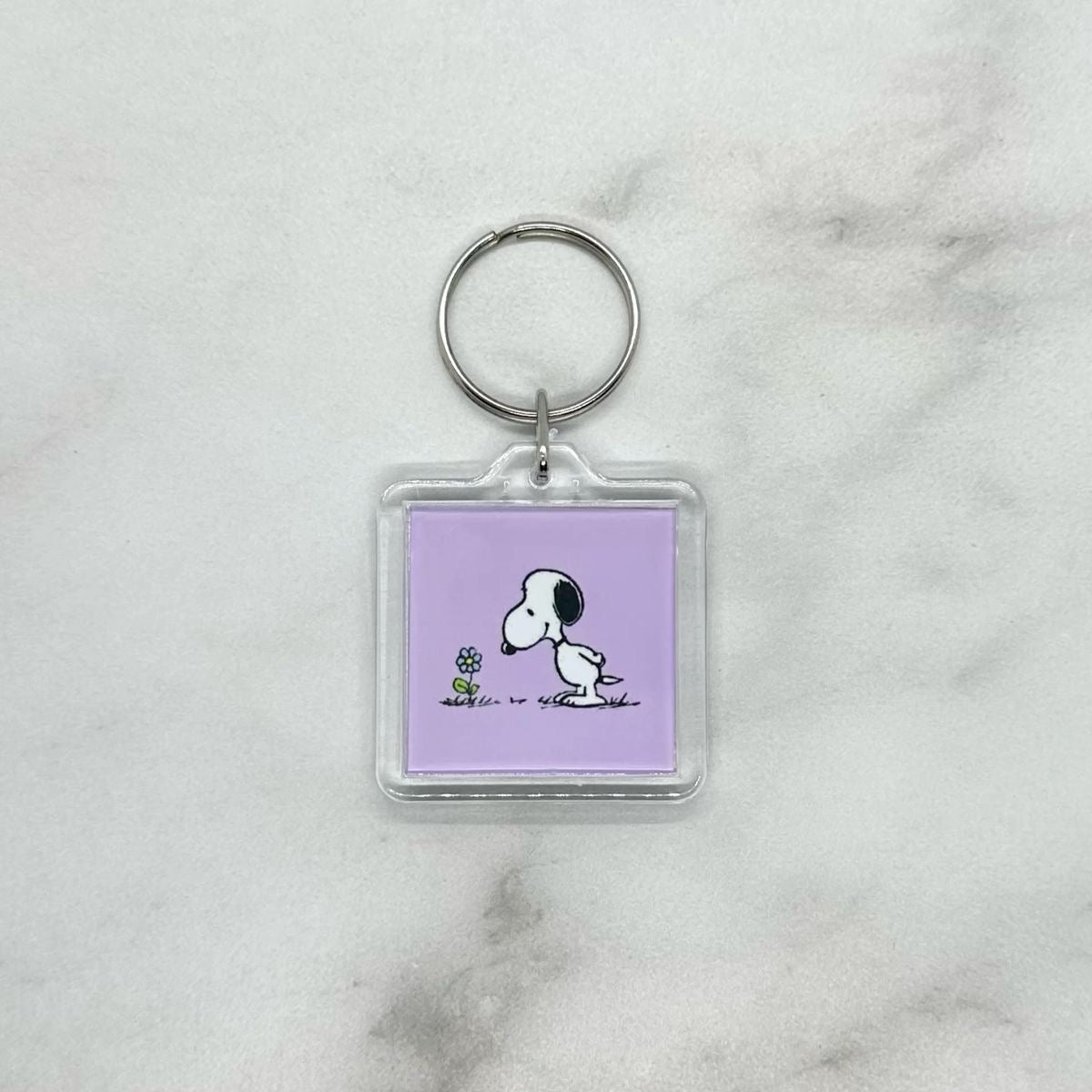Cute acrylic photo keychain of Snoopy smelling a flower. Purple background.